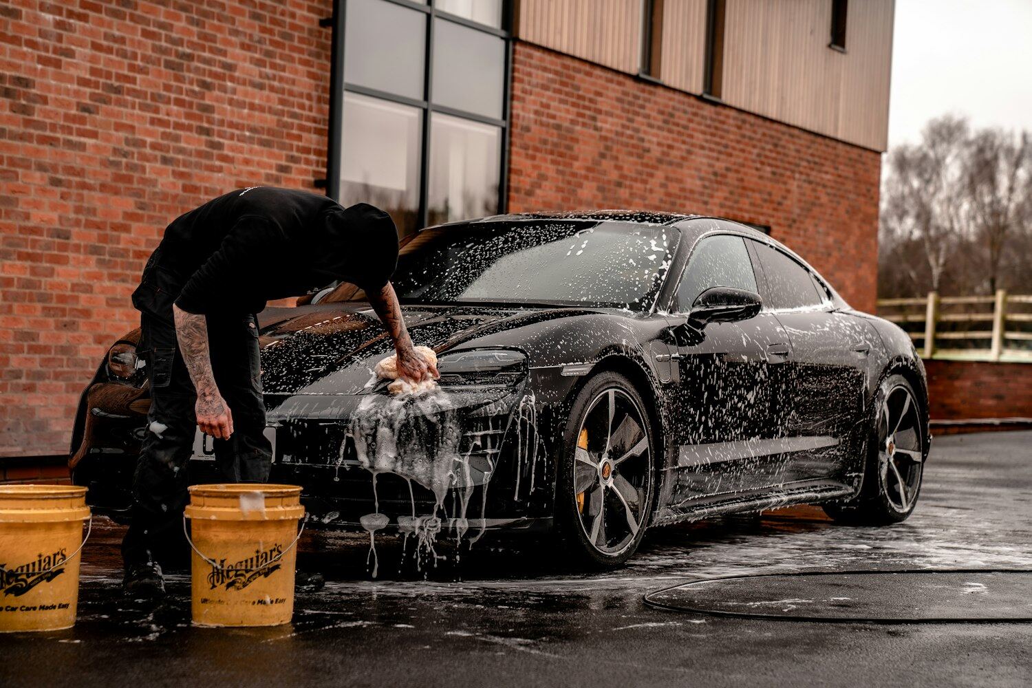 A detailer washing a dark sedan on location, with buckets on the ground beside the vehicle.